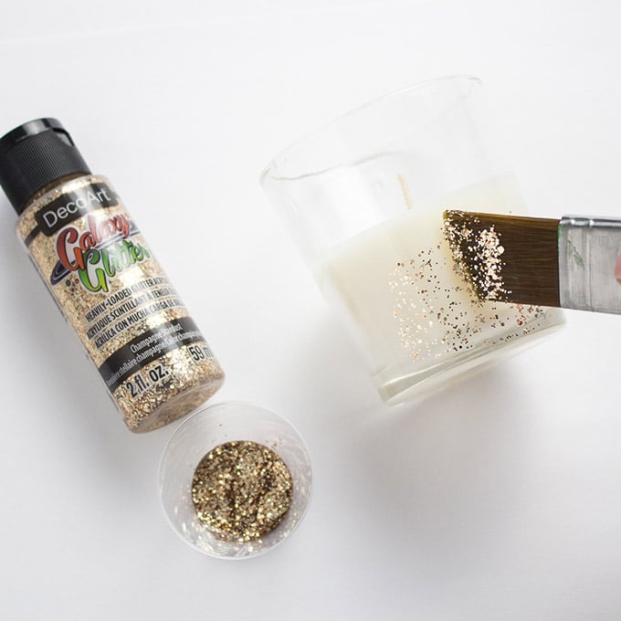 A bottle of Galaxy Glitter paint beside a glass votive candle holder which is being painted with the gold glitter.