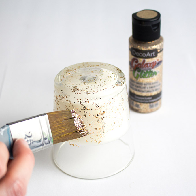 Applying gold glitter paint to the bottom half of a clear glass votive candle holder with a paint brush.