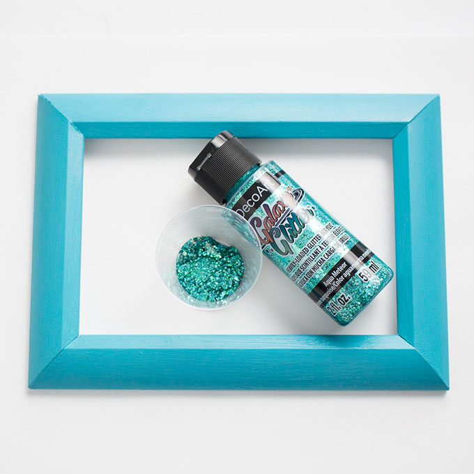 Bottle of aqua Galaxy glitter paint in the center of a 4x6 photo frame painted turquoise.