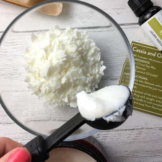 Adding a scoop of solid coconut oil to a clear glass bowl filled with soy wax flakes to make DIY fall candles.