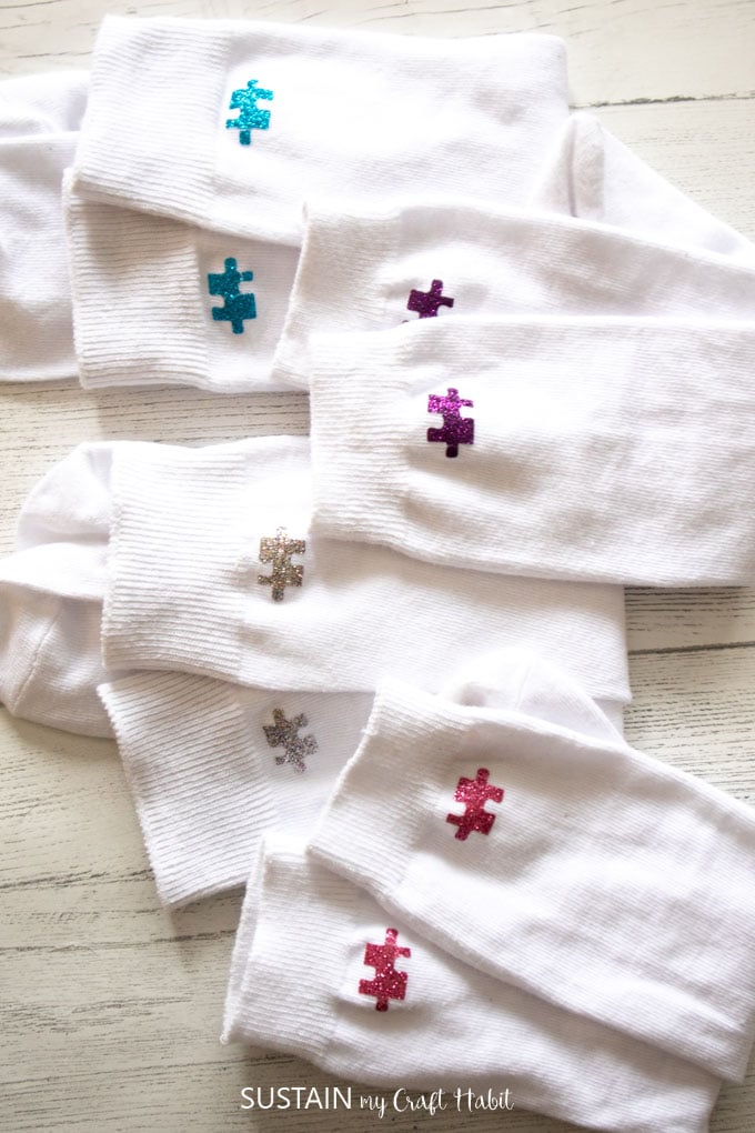 White socks lined up on a white wooden surface with different colors of puzzle pieces adhered with Cricut iron-on glitter vinyl.