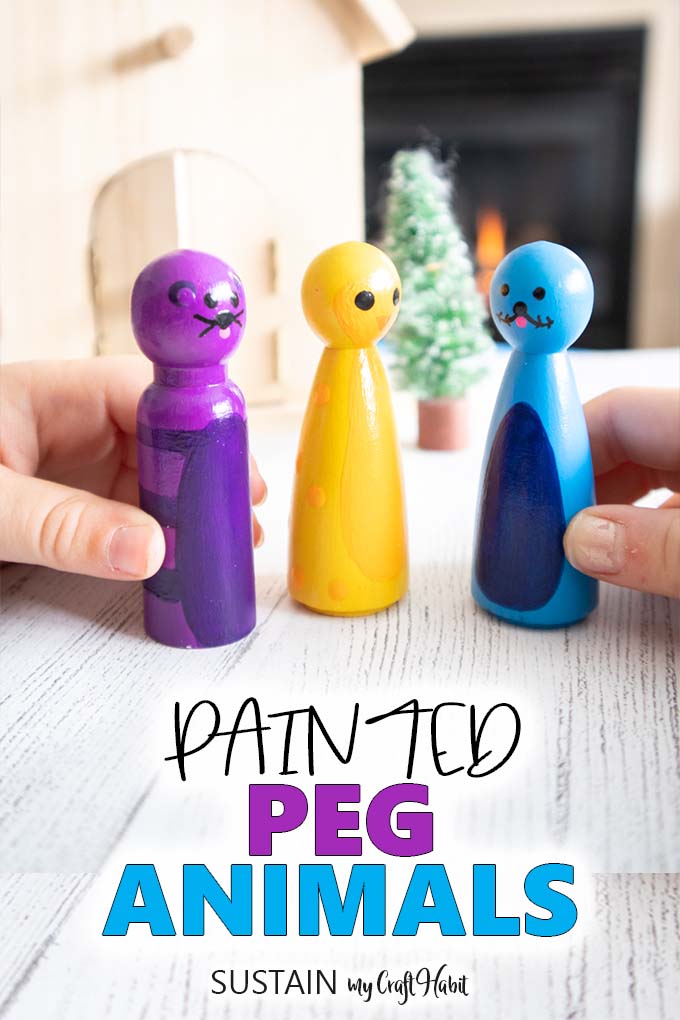 Image of hands holding wooden pegs painted as a purple polar bear, blue seal and yellow penguin. Also with the words "Painted peg animals" placed underneath.