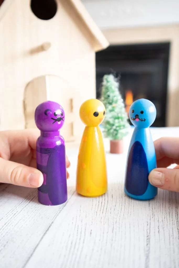 Two hand holding wooden pegs painted as a purple polar bear, blue seal and yellow penguin.