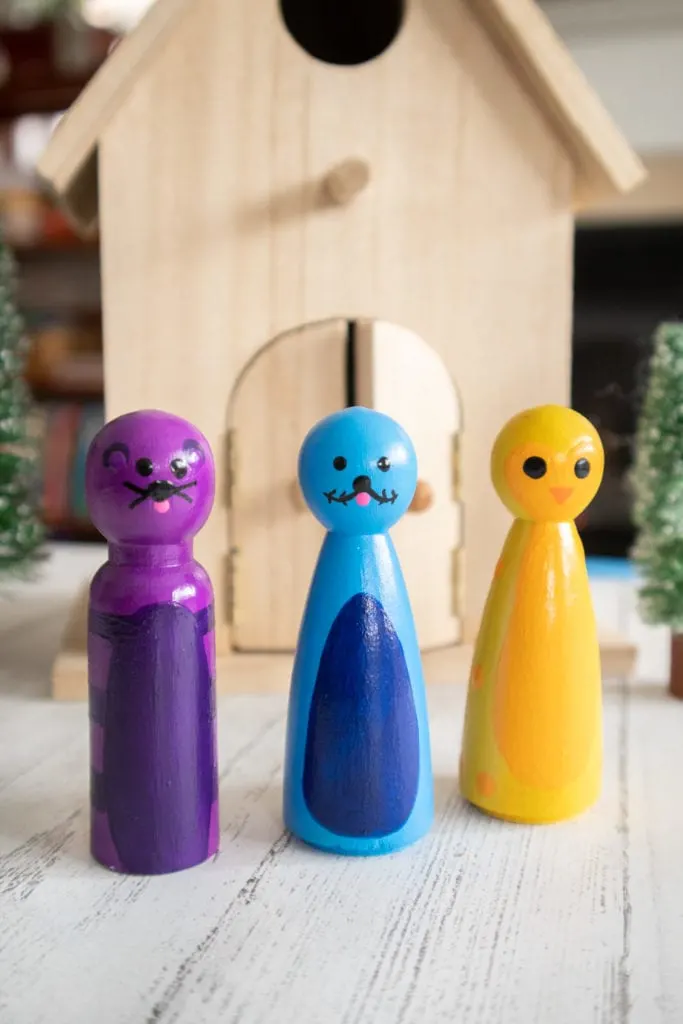 Wooden pegs painted as a purple polar bear, blue seal and yellow penguin arranged side by side in front of an unfinished wood bird house.