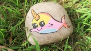 Rainbow narwhal painted on a rock and placed in the grass.
