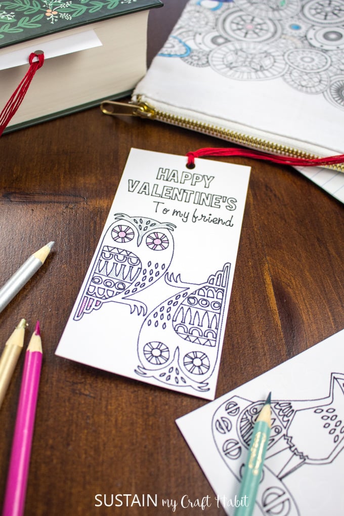 A Happy Valentine's bookmark with two owl images meant to be colored, on a dark wooden surface surrounded by pencil crayons.