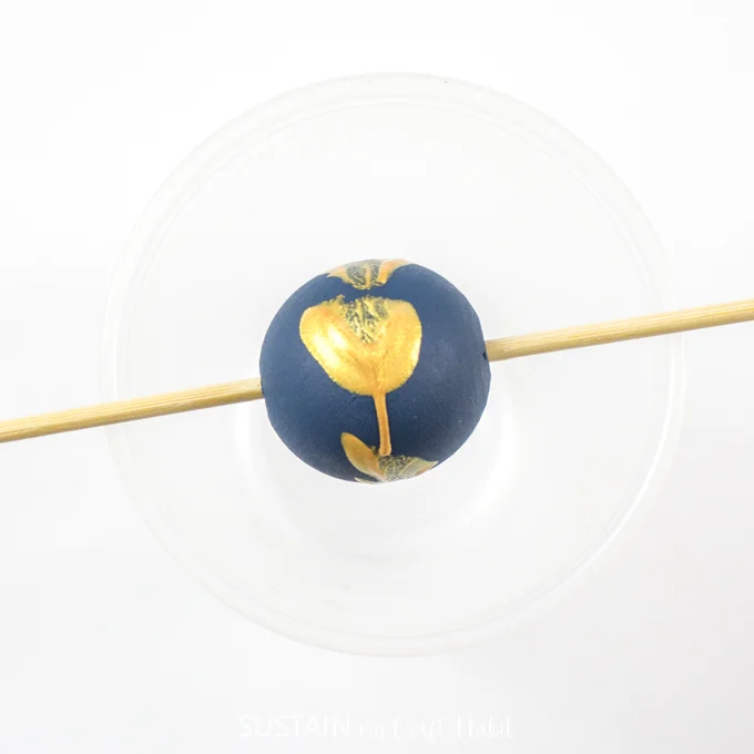 Blue painted wood bead with gold splatter placed on wooden skewer.