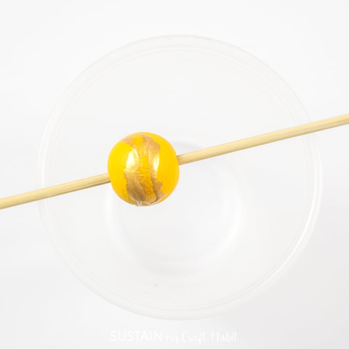 Yellow painted bead with gold splatter pattern placed on a wooden skewer.