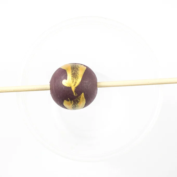 Purple painted wood bead with gold splatter placed on a wooden skewer.