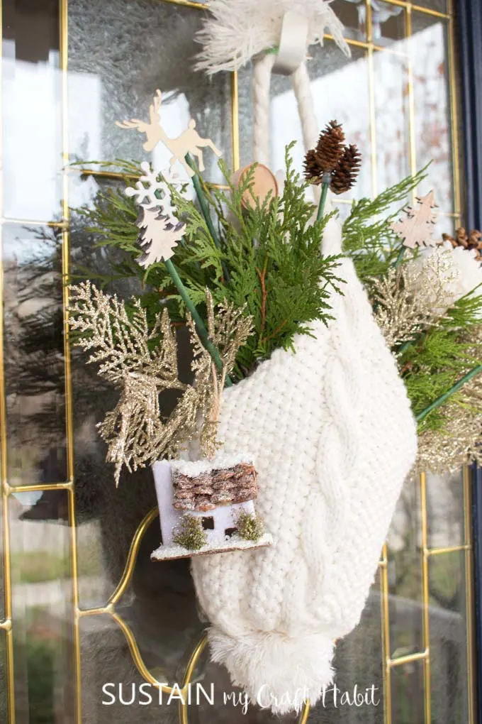 A white winter hat hanging from a glass door. The hat is filled with greenery and small decorative picks such as a snowflakes, trees, pine cones, wood slices and a birdhouse.