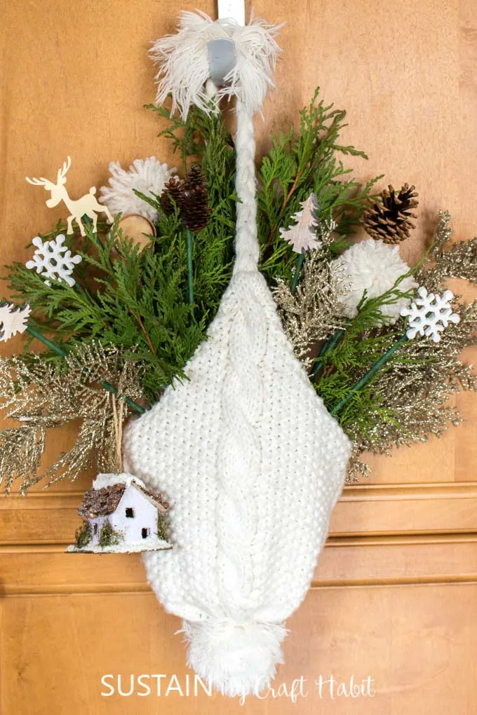 A white winter hat hanging from a hook. The hat is filled with greenery and small decorative picks such as a snowflakes, trees, pine cones, wood slices and a birdhouse.