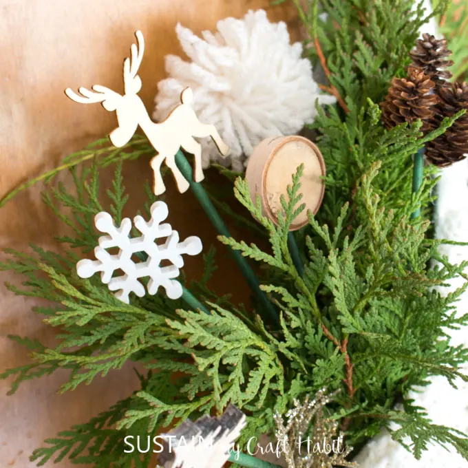 A close up of greenery mixed with decorative picks such as a snowflake, a reindeer,wood slice, a pom pom and pine cones.