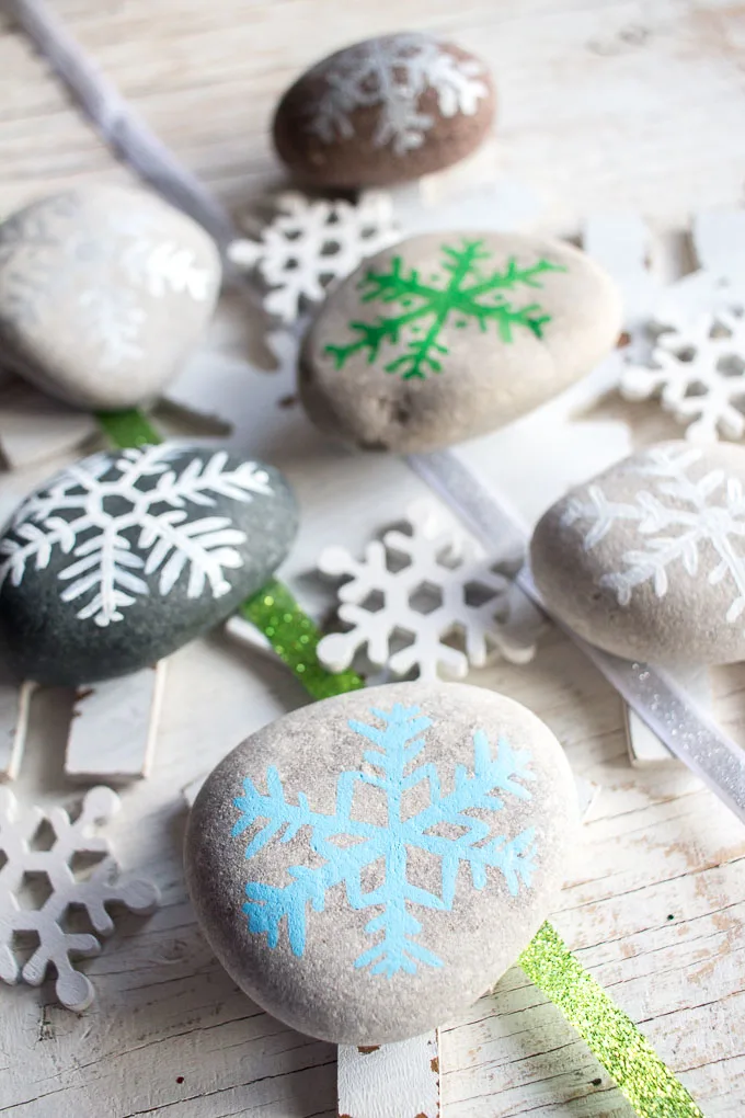 Six rocks painted with snow flake designs. They're placed on top of snowflake decorations and a white table.