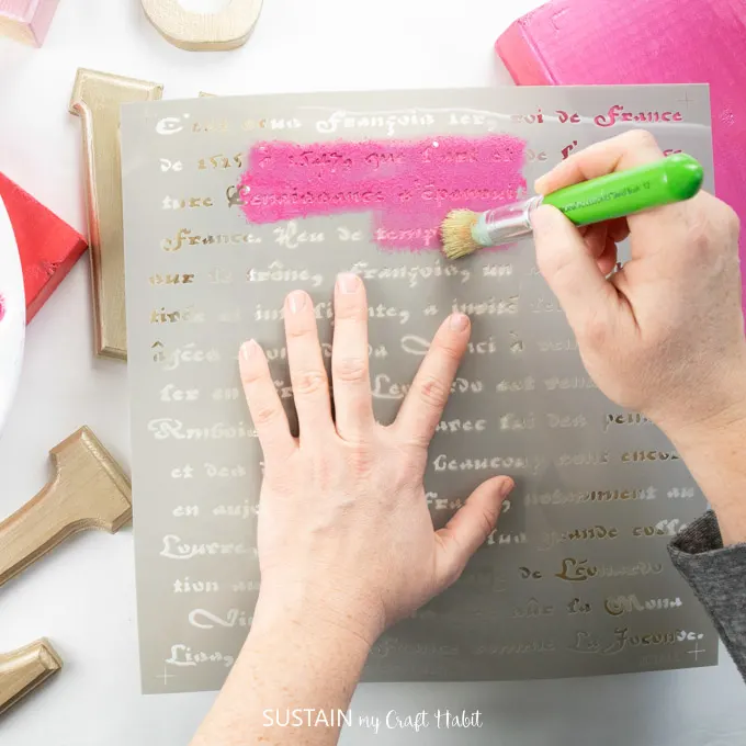 Painting over a french script stencil with pink paint onto a wooden  block.