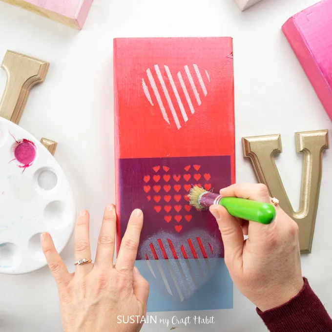 Painting over a heart stencil with silver paint onto a red painted wooden block.