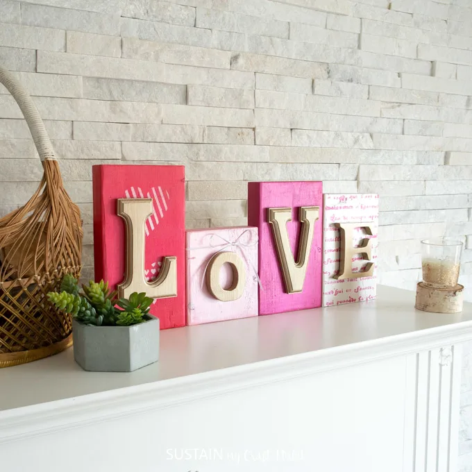 Colorful wood blocks with gold letters that spell out "love". They're sitting on a mantel behind a green succulent plant and candle.