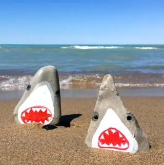 Two shark painted rocks on the beach with the lake in behind.