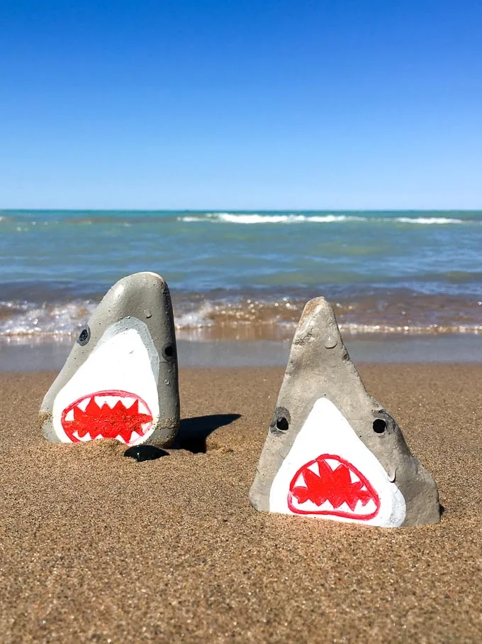 Two shark painted rocks on the beach with the lake in behind.