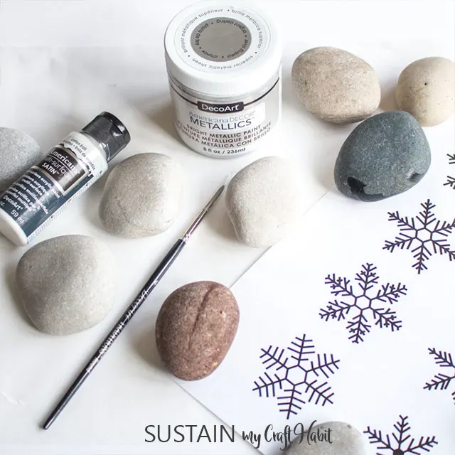 Supplies needed for the rock painting. Includes eight rocks, a paint brush, paint and a snowflake template.