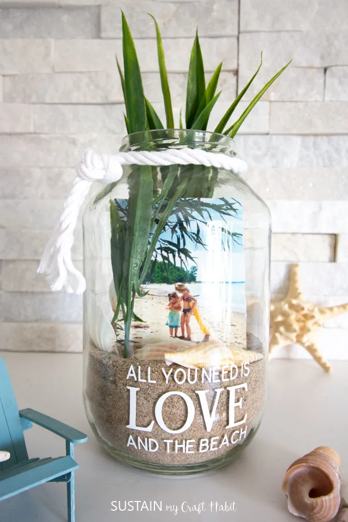 Clear jar filled with sand, sea shells, a picture and long faux greenery. The jar has white rope tied around the rim and is placed near a starfish, sea shell and Adirondack chair.