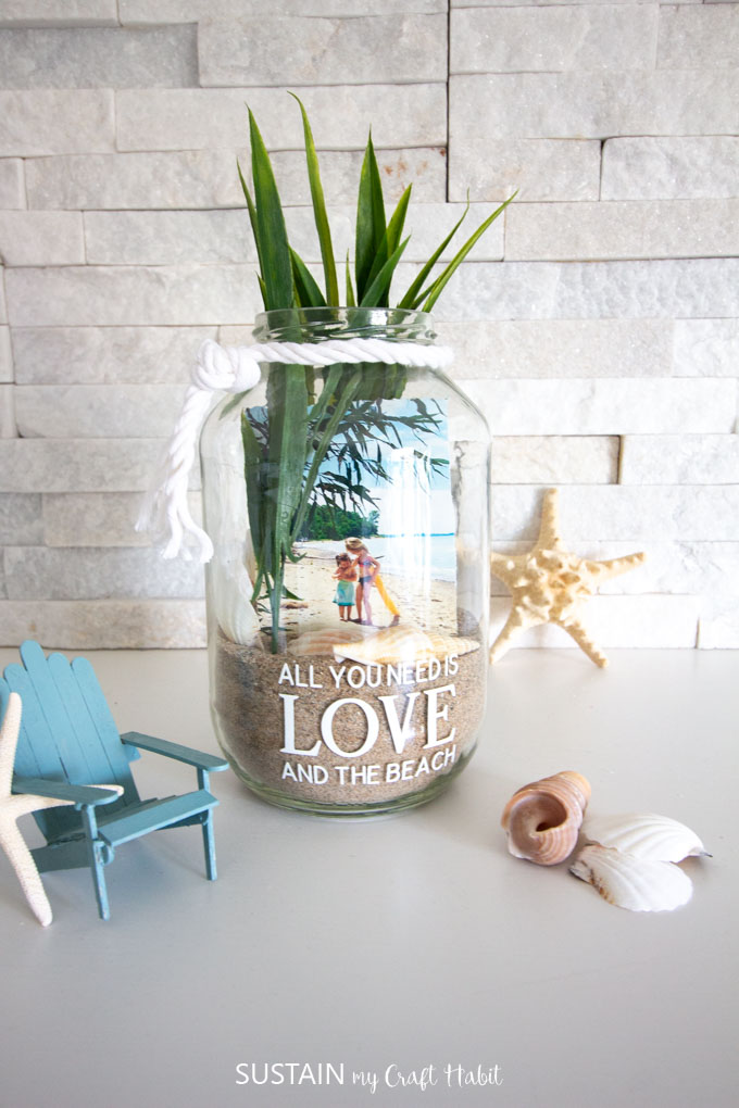 Finished project. Clear jar filled with sand, sea shells, a picture and long faux greenery. The jar has white rope tied around the rim and is placed near a starfish, sea shell and Adirondack chair.