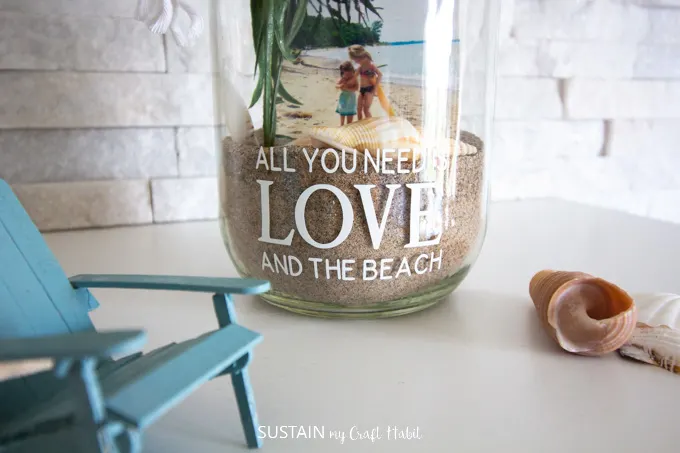 Showing the bottom of the glass jar with the cut out phrase, sand, sea shells and pictures.