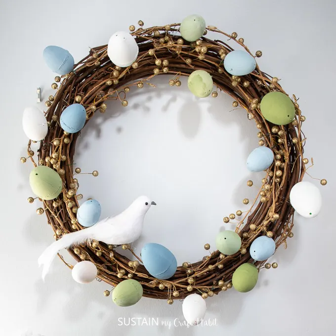 Grapevine wreath decorated with painted Easter eggs, gold garland and a decorative bird 