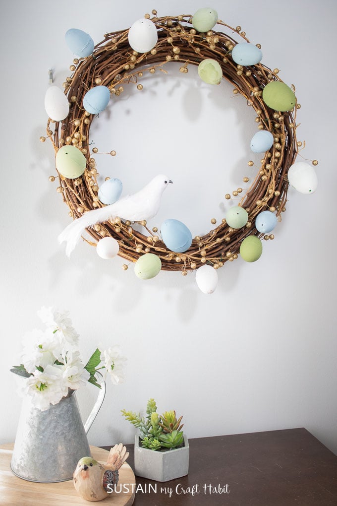 Grapevine wreath decorated with painted Easter eggs, gold garland and a decorative bird hung on the wall over a table with a decorative bird, succulents and a milk canister with flowers.