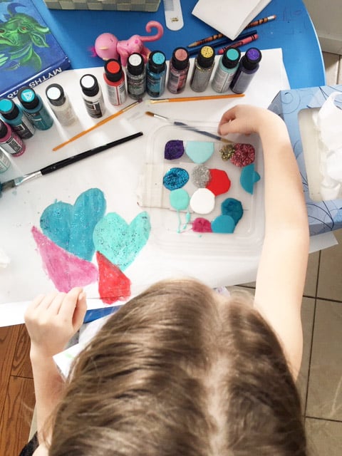 Child sitting at table with a row of paints in front of her and painting a picture.