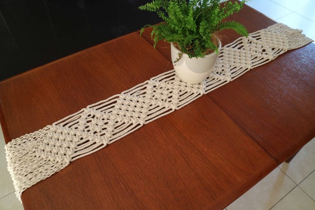 A completed macrame table runner on a cherry colored wood table surface.