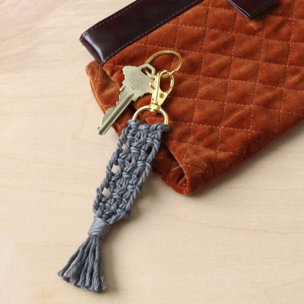 Close up image of a gray macrame keychain connected to a gold clasp resting against a rust colored purse.