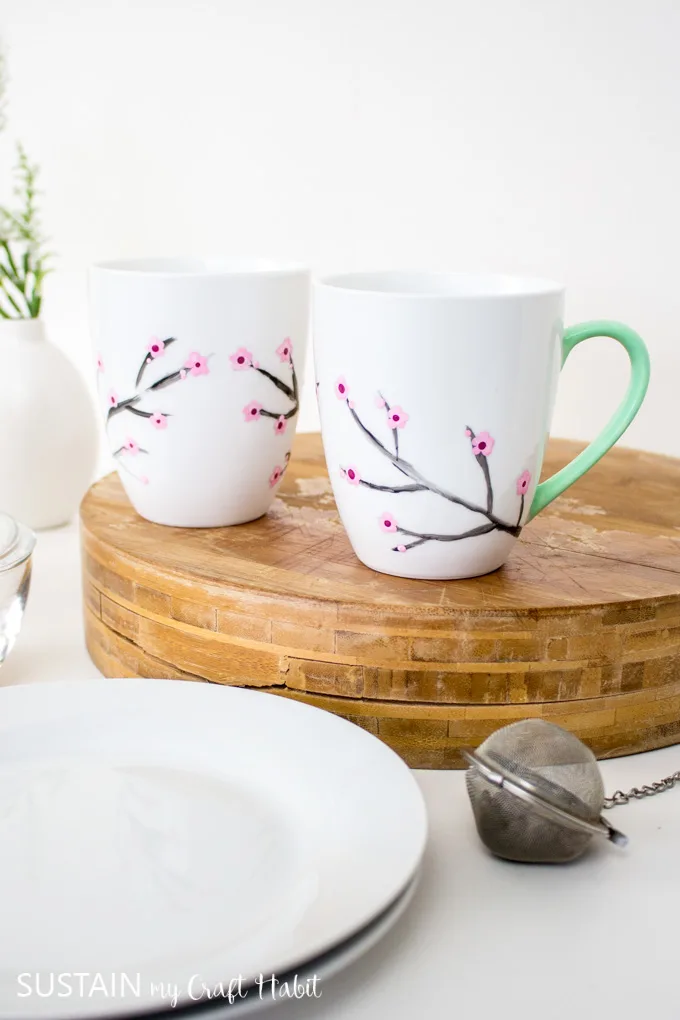 Two white mugs painted with cherry blossoms and green handles. The mugs are sitting on a wood tray beside white plates, a vase and tea infuser.