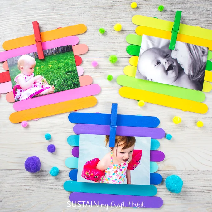 Colorfully painted Popsicle sticks made into photo frames holding a picture in each frame by a painted clothes pin. The frames are surrounded by pompoms.