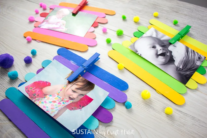 Colorfully painted Popsicle sticks made into photo frames holding a picture in each frame by a painted clothes pin. The frames are surrounded by pompoms and buttons.