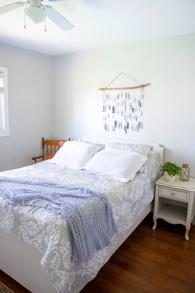 Wall decor paper feathers and driftwood hanging in a bedroom wall above a bed, next to a side table and wood chair.