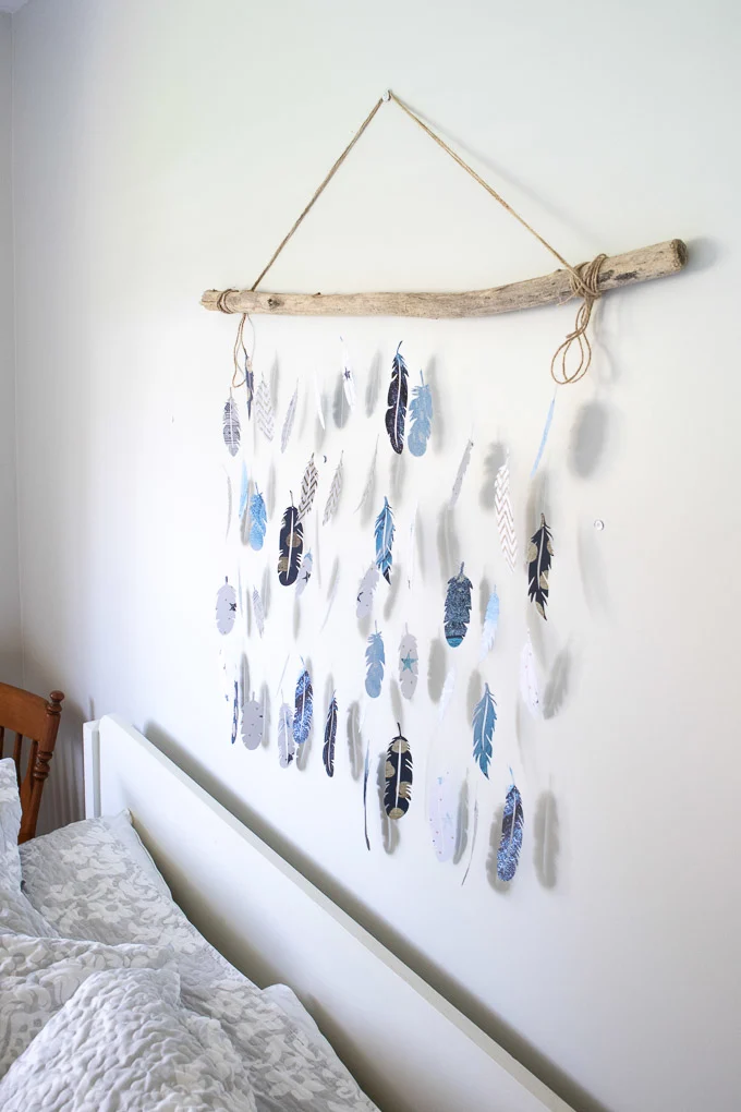 Wall decor made with blue, white and patterned paper feathers hanging from a piece of driftwood by a fishing line.