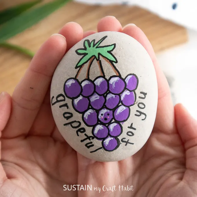 Holding the finished rock painted with grapes and text "grapeful for you."