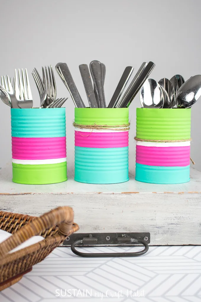Colorful and painted tin cans upcycled into a diy utensil holder filled with silver forks, knives and spoons.