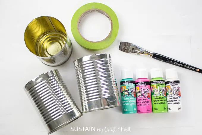 Materials needed to make a colorful utensil holder, including tin cans, paint, tape and a paint brush.