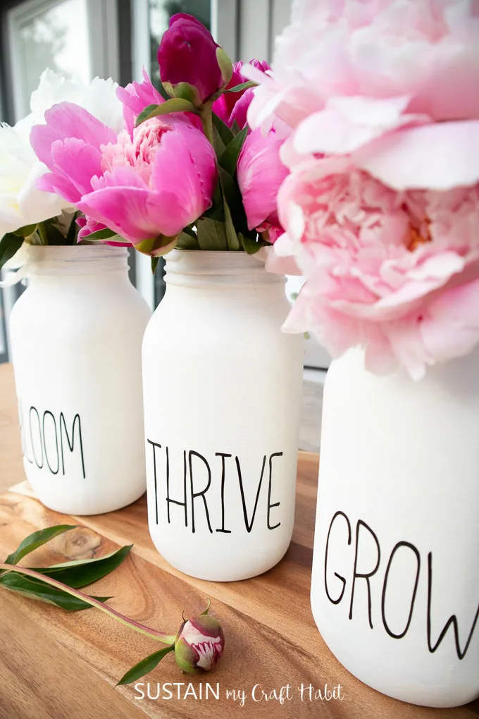 Finished chalky painted mason jar flower vases with words on the front saying "bloom," "thrive," "grow" and filled with peony flowers.