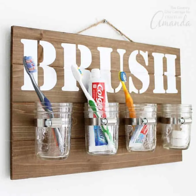 A wood board with four mason jars affixed and filled with tooth brushes and tooth paste. The wood sign has the word brush stencilled on it in white paint.