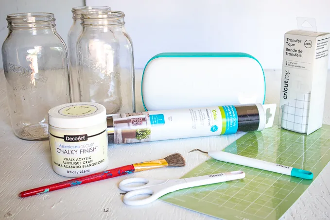 Materials needed to make a chalky painted mason jar glower vase, including mason jars, paint, paintbrush, Cricut machine, vinyl, a grip mat and Cricut weeding tools.