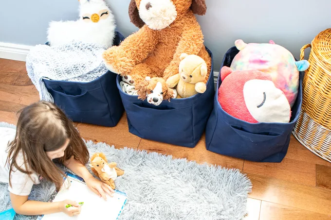 A small girl drawing pictures in front of three fabric storage cubes filled with toys.