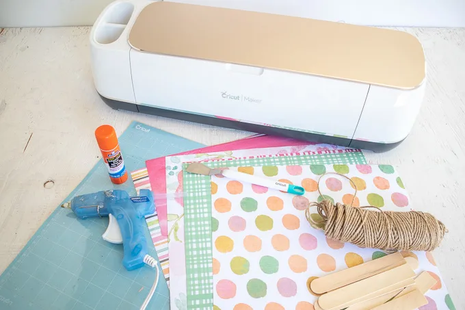 Materials needed to make a paper popsicle garland, including a Cricut machine, hot glue gun, glue stick, twine, popsicle sticks and colorful paper.