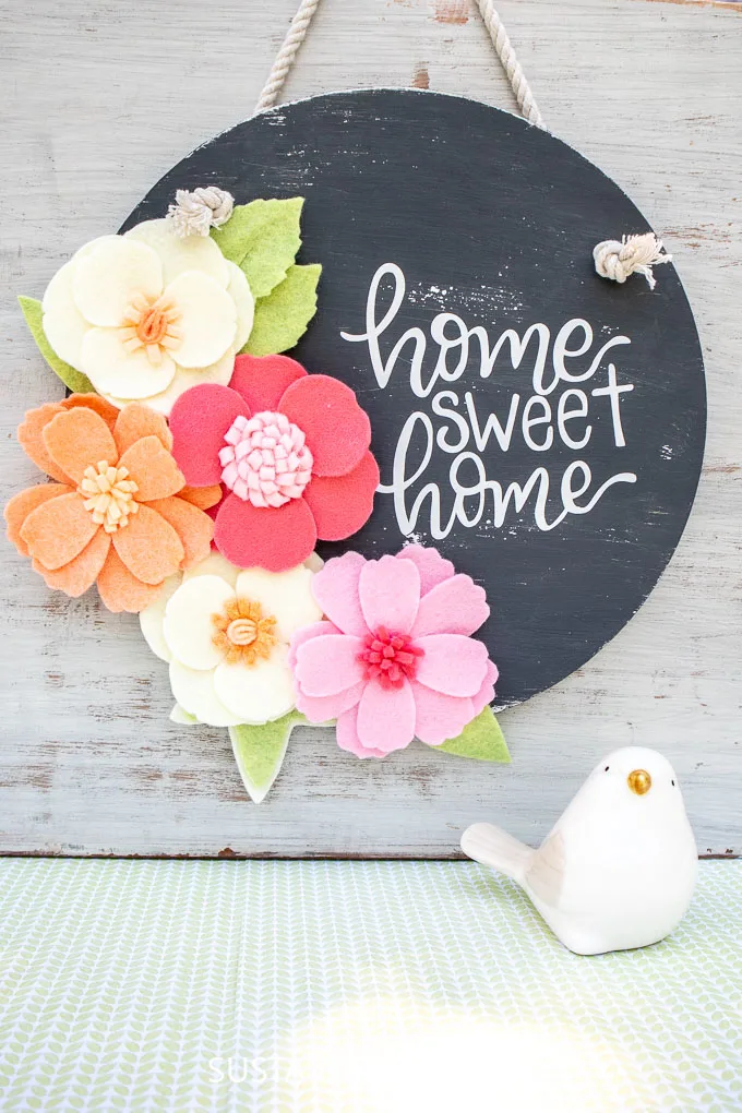 A round dark gray painted wood sign with colorful felt flowers on one side and the handlettered phrase "home sweet home" on the other.