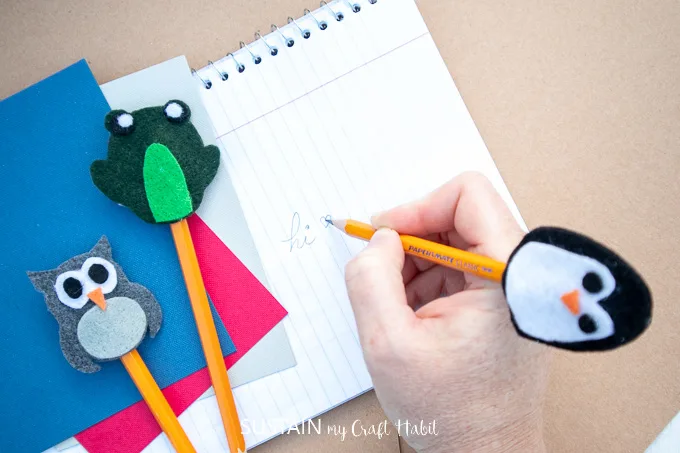 Writing with a pencil that has a felt fabric penguin topper and next to frog and owl pencil toppers.