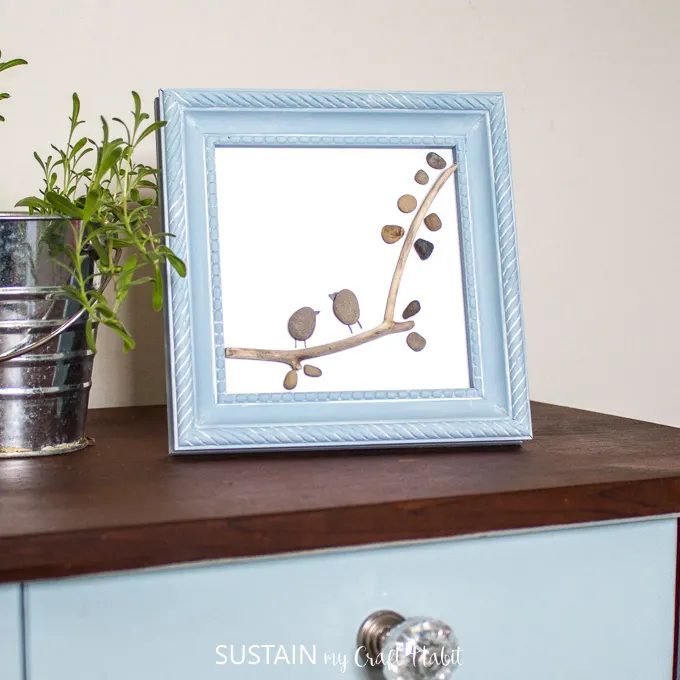 Completed pebble art in a small blue frame on a dark wood surface. A mall plotted plant is to the left side.