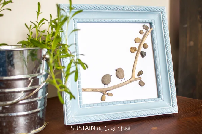 Close up of a blue frame pebble art resembling two birds perched on a tree branch.