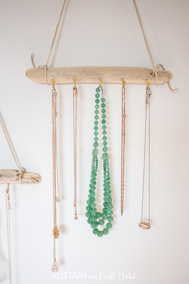 A driftwood jewelry hanger with gold hooks hanging on a wall holding various necklaces as an example of a DIY jewelry display idea.