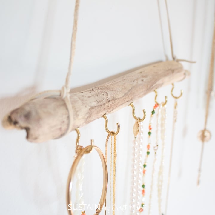 Homemade jewellery display idea with a long piece of driftwood and gold cup hooks. Various necklaces and bracelets hang on the hooks.